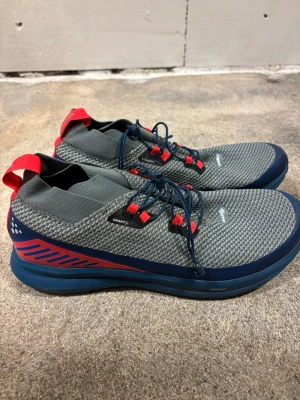 Grå sneakers från Craft med röda detaljer - Snygga grå sneakers från Craft med stickad ovandel, blå sula och röda detaljer vid snörningen och hälen. Skorna har rund tå, platt sula och snörning för bra passform. Perfekta för dig som gillar sportig stil.