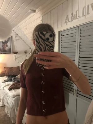 Vinröd topp med rosettdetaljer Zara - Vinröd croppad topp från Zara med korta ärmar och dekorativa rosettformade knappar framtill. Rund halsringning och mjukt stickat material ger en trendig och bekväm look. Perfekt för dig som gillar söta detaljer och vill sticka ut. Aldrig använd