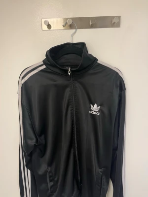  Svart Adidas track jacket med dragkedja storlek S och M passar denna tröja  - Denna tröja passar folk med storlek S och M. Old school adidas. Klassisk svart Adidas track jacket med vita ränder längs ärmarna och dragkedja framtill. Tröjan har hög krage, två fickor och Adidas Trefoil-logga på bröstet och ryggen. Perfekt för en sportig och avslappnad stil.