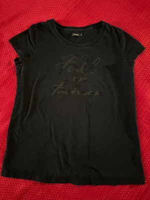 Svart t-shirt från O'STIN med text - Svart t-shirt från O'STIN med texten 'Fall in Fashion' framtill i glittriga detaljer. Klassisk rund halsringning och korta ärmar. Perfekt för en stilren och enkel look.