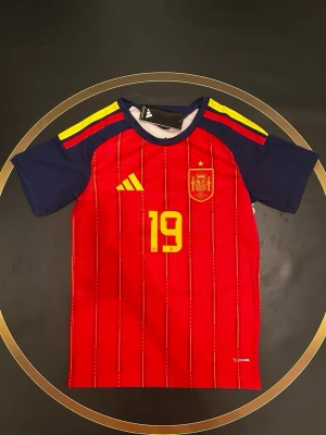 Spaniens matchtröja Adidas röd - Röd matchtröja från Adidas för Spaniens landslag med gula detaljer, nummer 19 på bröstet och landslagets emblem. Tröjan har korta ärmar i mörkblått och gula ränder på axlarna. Perfekt för fotbollsfans som vill visa sitt stöd. S och L