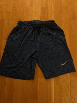 Mörkblå träningsshorts från Nike - Mörkblå träningsshorts från Nike med elastisk midja och snörning. Shortsen har en diskret melerad look och klassisk Nike-logga på ena benet. Perfekta för sport eller chill dagar.