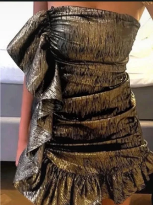 Guldig metallic one shoulder klänning - Säljer en unik guldig kortklänning med metallic-look och stora volangdetaljer. Klänningen är axelbandslös med en dramatisk oneshoulder-design och har en figurnära passform. Perfekt för dig som vill sticka ut och glänsa. Liten i storleken!!