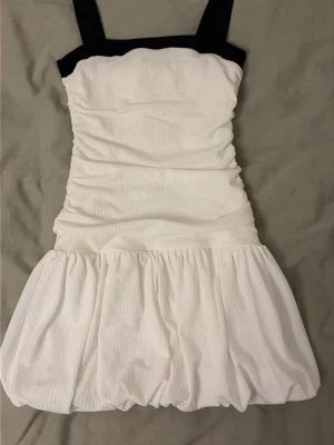 Vit kort klänning med svarta band - Supersöt vit kort klänning med svarta axelband och fyrkantig urringning. Klänningen har en tight smockad överdel och en voluminös kjol med ballongformad nederdel