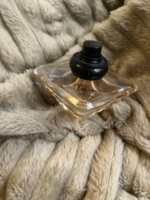 Lancôme  parfym - Säljer en elegant parfym från Lancôme, modellen Trésor. Flaskan är fyrkantig med mjuka kanter och har en svart rund kork. Glaset är transparent med en svag persikofärgad ton på vätskan. Perfekt för dig som gillar klassiska och lyxiga dofter.