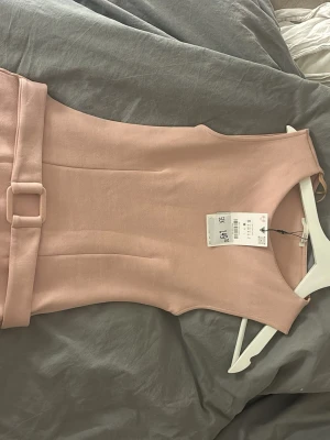 Ljusrosa klänning med bälte från Vero Moda - Snygg ljusrosa kortklänning från Zara med matchande bälte i midjan. Klänningen är ärmlös och har en figurnära passform som framhäver siluetten. Tillverkad i ett mjukt material som känns bekvämt mot huden. Perfekt för dig som gillar stilrena och feminina plagg.