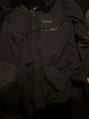 Svart ziptröja från BOSS - Snygg svart ziptröja från BOSS med grön logga på bröstet. Tröjan har hel dragkedja framtill, långa ärmar och diskreta detaljer. Perfekt för en avslappnad och stilren look.