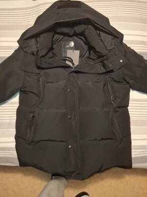 Svart pufferjacka från Canada Goose - Svart pufferjacka från Canada Goose med huva, dragkedja och knappar framtill. Jackan har quiltad design, stora fickor med dragkedja och den klassiska loggan på ärmen. Tillverkad i slitstark polyester och perfekt för kalla vinterdagar.