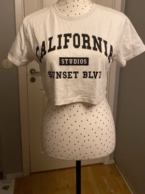 Vit croppad t-shirt California - Snygg vit croppad t-shirt med trycket 'California Studios Sunset Blvd' i svart. T-shirten har rund halsringning och korta ärmar, perfekt för en avslappnad och trendig stil.