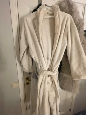 Krämvit fluffig morgonrock S/M - Mysig och fluffig morgonrock i krämvit färg med skärp i midjan och stora fickor. Tillverkad i mjukt frottéliknande material som känns skönt mot huden. Perfekt för lata morgnar eller kvällar hemma.