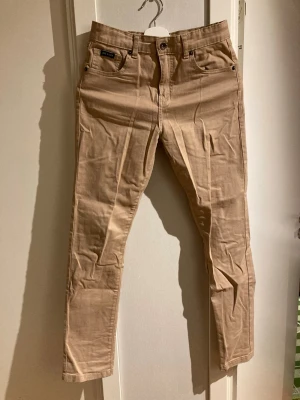 Beige chinos från Lyle & Scott - Snygga beige chinos från Lyle & Scott med klassisk femficksdesign och knappgylf. Byxorna har en rak passform och är tillverkade i mjuk bomull som ger en skön känsla. Perfekta för dig som vill ha en clean och stilren look.