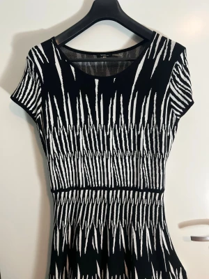 Svartvit mönstrad klänning från Vero Moda - Säljer en svartvit klänning från Vero Moda med grafiskt mönster. Klänningen har rund halsringning, korta ärmar och markerad midja. Materialet är stretchigt och bekvämt, vilket ger en snygg passform. Perfekt för dig som gillar statement-plagg!