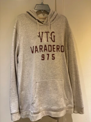 Beige hoodie Jack and Jones  - Beige/vit hoodie med huva och dragsko, stor magficka och tryck i vinrött framtill med texten 'VTG VARADERO 975'. Mjuk bomullsblandning och avslappnad passform, perfekt för en chill stil.