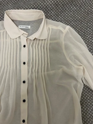 Beige transparent blus med svarta knappar - Stilren beige blus från Second Female i transparent material. Blusen har trekvarts ärmar, klassisk krage och svarta knappar framtill. Dekorativa veck på bröstet ger en snygg detalj. Perfekt att styla lager på lager.