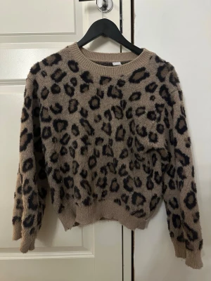 Leopardmönstrad stickad tröja - Säljer en fluffig stickad tröja med leopardmönster i beige och svart. Tröjan har rund halsringning, långa ärmar och ribbade muddar. Perfekt för dig som vill ha en trendig och mysig look med djurmönster. Den har ett litet hål men ingenting som syns tydligt. Storlek 146/162=xxs/xs
