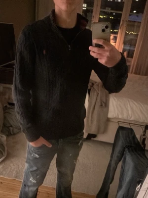 Mörkblå kabelstickad tröja Polo Ralph Lauren - Mörkblå kabelstickad tröja från Polo Ralph Lauren med halv dragkedja och röd logga på bröstet. Tröjan har ribbade muddar och krage med röda ränder på insidan. Perfekt för dig som gillar klassisk och stilren design.