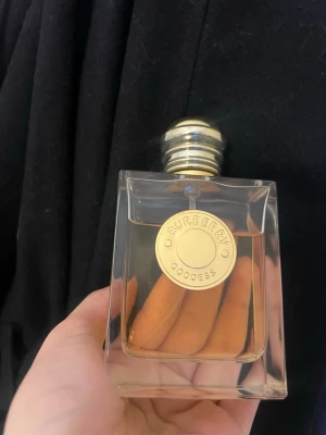 Burberry Goddess parfym 100ml - Burberry Goddess parfym i en stilren, fyrkantig glasflaska med guldlock och rund gulddetalj framtill. Doften har en elegant och lyxig känsla, perfekt för dig som vill ha något exklusivt. Flaskan är transparent och visar den ljusgula vätskan inuti. Cirka 70% kvar 