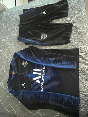 PSG träningsset svart/blå Puma - Säljer ett PSG träningsset från Puma i svart med blå detaljer. Setet består av långärmad tröja och byxor, båda med PSG-logga och Puma-logga. Materialet är troligtvis polyester, perfekt för träning eller fotboll. Snygg design med vita och blå tryck på bröstet.