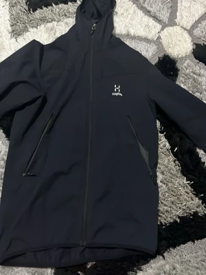 Svart vindjacka från Haglöfs med huva - Svart vindjacka från Haglöfs med huva och hel dragkedja framtill. Jackan har två snedställda fickor med dragkedja och diskret logga på bröstet. Perfekt för blåsiga dagar och outdoor-aktiviteter.