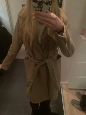 Beige omlottkappa med bälte - Snygg beige kappa med omlottmodell och bälte i midjan. Kappan har stora fickor framtill och en klassisk krage. Perfekt för dig som gillar stilrena och tidlösa plagg. Passar bra till både jeans och klänning.