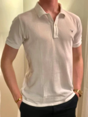 Vit pikétröja från GANT - Klassisk vit pikétröja från GANT med korta ärmar och krage. Tröjan har knappar framtill och den ikoniska GANT-loggan broderad på bröstet. Tillverkad i mjuk bomull för en bekväm och stilren look.