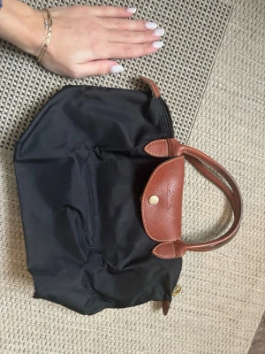 Svart handväska från Longchamp - Klassisk svart handväska från Longchamp med bruna skinnhandtag och lock. Väskan har guldfärgade metalldetaljer och dragkedja med Longchamp-logga. Perfekt storlek för vardagens essentials och enkel att matcha till olika outfits.