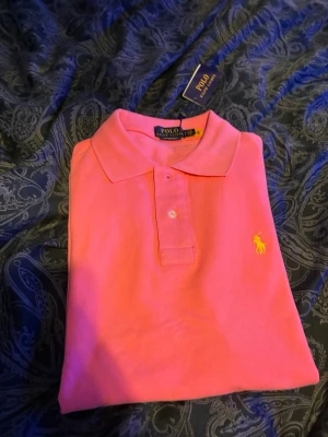 Rosa pikétröja från Polo Ralph Lauren - Snygg rosa pikétröja från Polo Ralph Lauren i modellen Custom Slim Fit. Tröjan har klassisk krage, knappar framtill och den ikoniska gula loggan broderad på bröstet. Perfekt för en stilren och sportig look.