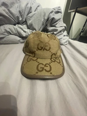 Beige keps från Gucci med GG-mönster - Säljer en beige keps från Gucci med det klassiska GG-mönstret i brunt över hela kepsen. Kepsen har böjd skärm och är tillverkad i canvasmaterial med detaljer i brunt läder runt skärmen. Perfekt för dig som vill ha en stilren och lyxig accessoar.