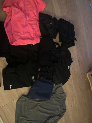 Träningskläder Nike och mixade märken - Säljer ett paket med träningskläder, inklusive ett par rosa shorts, flera svarta träningsplagg och en grå t-shirt. Några plagg är från Nike och andra från olika märken. Perfekt för gymmet eller löpning. Materialen är funktionsmaterial som andas.