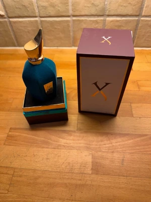 Xerjoff blå parfymflaska med låda ANVÄND LITE  - Lyxig parfym från Xerjoff i en unik blå sammetsklädd flaska med guldfärgad metallkork och gulddetaljer. Flaskan har en elegant, rundad form och levereras i en exklusiv ask med matchande design och Xerjoff-logga på både flaska och låda. ANVÄND LITE.