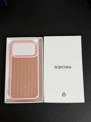 Rosa Iphone 17 pro max Rimowa mobilskal - Snyggt mobilskal från Rimowa i rosa och kopparfärgad aluminium med klassisk räfflad design. Skalets ram är i rosa plast och baksidan har en metallisk finish. Passar perfekt för dig som vill ha ett stilrent och tåligt skydd till din iPhone.