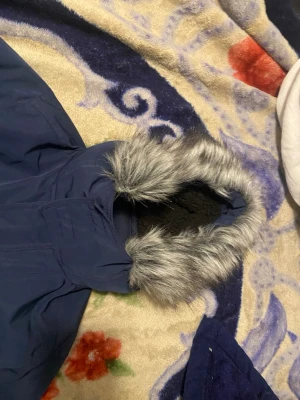 Blå parkas med fuskpäls från Hollister - Snygg mörkblå parkas från Hollister med huva kantad av grå fuskpäls. Jackan är en del av Hollister's All-Weather Collection och har ett mjukt, svart innerfoder. Perfekt för kyliga dagar och har en klassisk parkas-look.