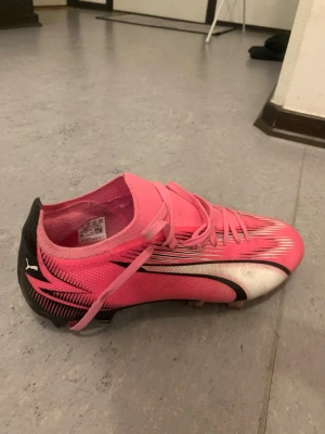 Puma rosa fotbollsskor med snörning - Snygga fotbollsskor från Puma i rosa, svart och vitt. Skorna har snörning, låg profil och mönstrad ovandel i syntetmaterial. Yttersulan har fasta dobbar för bra grepp på planen. Perfekta för dig som vill sticka ut på matchen!