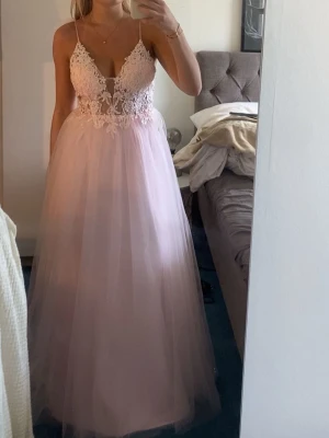 Ljusrosa långklänning med spetsdetaljer - Romantisk ljusrosa långklänning med tunn tyllkjol och vacker spets upptill. Klänningen har smala axelband, djup v-ringning och broderade detaljer över bysten. Perfekt för speciella tillfällen.