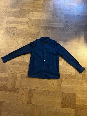 Mörkblå skjorta med knappar från Gant - Stilren mörkblå skjorta med klassisk krage och vita knappar framtill från Gant. Skjortan har lång ärm och en liten bröstficka med diskret logga. Perfekt till jeans eller chinos för en clean look.