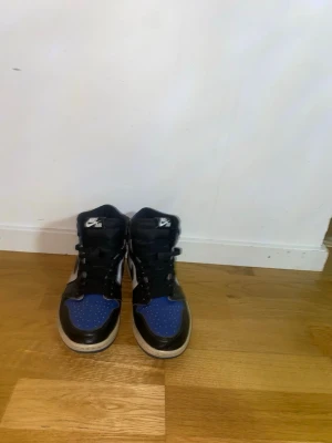 Nike Air Jordan 1 svart/blå sneakers - Säljer ett par Nike Air Jordan 1 sneakers i svart, blått och vitt. Klassisk high-top modell med snörning och rund tå. Ovandelen är i skinn och har den ikoniska Nike-loggan på plösen. Perfekta för dig som gillar streetwear och vill sticka ut.