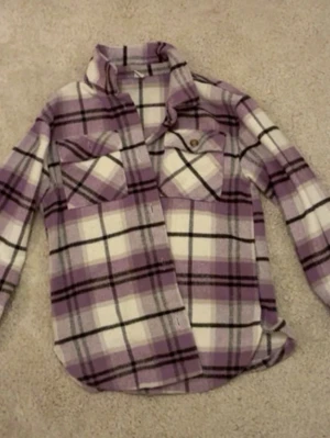 Lila och vit rutig overshirt - Overshirt i lila, vit och svart rutig design med två bröstfickor och knappar framtill. Skön och avslappnad passform, perfekt att slänga över  en t-shirt. Långärmad och tillverkad i mjukt bomullsmaterial.