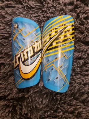Nike Mercurial benskydd blå/gul - Säljer ett par Nike Mercurial benskydd i blått med gula och vita detaljer samt tillhörande blåa sleeves med gul logga. Snygg design med grafiskt mönster och stor Nike swoosh. Perfekta för fotbollsträning eller match. Priset kan diskuteras!