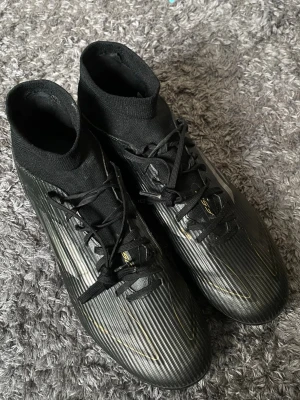 Svarta adidas fotbollsskor med snörning - Säljer ett par svarta adidas fotbollsskor med högt skaft och snörning. Skorna har tunna guldfärgade detaljer och är tillverkade i syntetmaterial för bra grepp och känsla på planen. Perfekta för dig som vill ha stilrena och funktionella skor till matchen.                        Helt nya, Pris kan självklart diskuteras 