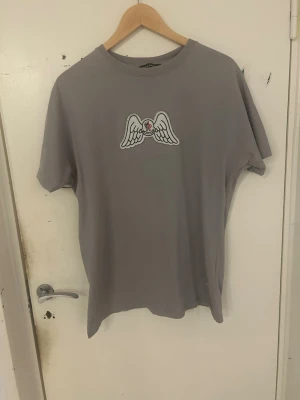 Grå t-shirt från Palm Angels - Grå t-shirt från Palm Angels med ett broderat motiv av vita änglavingar och ett rött hjärta på bröstet. Klassisk rund halsringning och avslappnad passform. Perfekt för dig som gillar streetwear och stilrena detaljer.