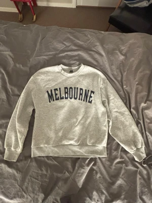 Grå sweatshirt med MELBOURNE-tryck - Snygg grå sweatshirt med svart text 'MELBOURNE' över bröstet. Klassisk rund halsringning och ribbade muddar vid ärmslut och nederkant. Perfekt för en avslappnad och trendig stil.