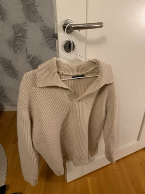 Beige stickad tröja med krage - Säljer en beige stickad tröja från j lindeberg med bred krage och v-ringning. Tröjan har en avslappnad passform och ribbstickat material som ger en trendig look. Perfekt att styla med jeans eller kjol för en snygg outfit. Nypris 2200