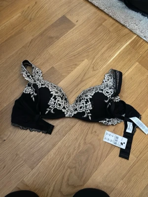 Svart spets-bh med broderi - Supersnygg svart bh från Intimissimi med vacker beige spets och broderade blommor. Bh:n har vadderade kupor och dekorativa spetsdetaljer på axelbanden. Perfekt för dig som gillar en lyxig och feminin look. Storlek 75B