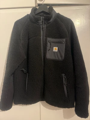 Svart teddyjacka från Carhartt WIP - Mysig svart teddyjacka från Carhartt WIP med hel dragkedja, hög krage och kontrastficka på bröstet med logga. Jackan har två sidofickor med dragkedja och är perfekt för kalla dagar. Tillverkad i mjuk fleece och har en clean, stilren look.