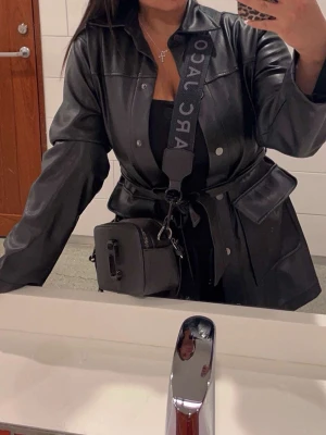 Svart skinnjacka med bälte - Snygg svart skinnjacka med bälte i midjan och klassisk krage. Jackan har långa ärmar, knappar framtill och stora fickor på sidorna. Perfekt för dig som vill ha en trendig och stilren look.