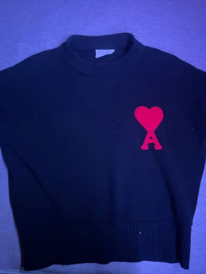 Svart stickad tröja från Ami Paris - Svart stickad tröja från Ami Paris med röd hjärtlogga och bokstaven A broderad på bröstet. Tröjan har ribbstickad hals, ärmslut och nederkant. Perfekt för dig som gillar stilrena plagg med en twist. OBS KAN SÄNKA PRIS