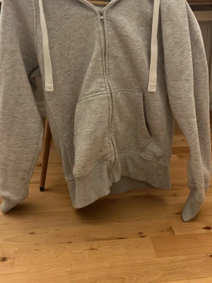 Russemerch tröja - En grå hoodie från russemerch med dragkedja och stora fickor framtill. Modellen har huva med breda snören och ribbade muddar vid ärmslut och nederkant. Perfekt för en avslappnad och stilren look.