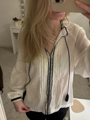 Vit blus med svarta detaljer och tofsar - Vit blus med svarta broderade detaljer längs knäppningen och kragen. Blusen har långa ärmar, v-ringning och dekorativa svarta knytband med tofsar. 