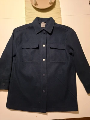Mörkblå overshirt från Primark - Stilren mörkblå overshirt från Primark med klassisk krage och tryckknappar framtill. Två stora bröstfickor med lock ger en cool look. Perfekt att bära som lager över en hoodie eller t-shirt. Materialet känns mjukt och har en ullkänsla.