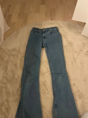Blå bootcut jeans med hög midja - Snygga blå bootcut jeans med hög midja och klassisk femficksdesign. Jeansen har en tidlös passform och är tillverkade i ett mjukt denimtyg som ger en skön känsla. Perfekta för dig som gillar en retroinspirerad look.